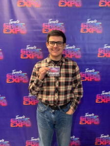 Dmitrii Afanasev standing at Toronto Game Expo holding a volunteer badge, smiling in front of the event's branded backdrop.